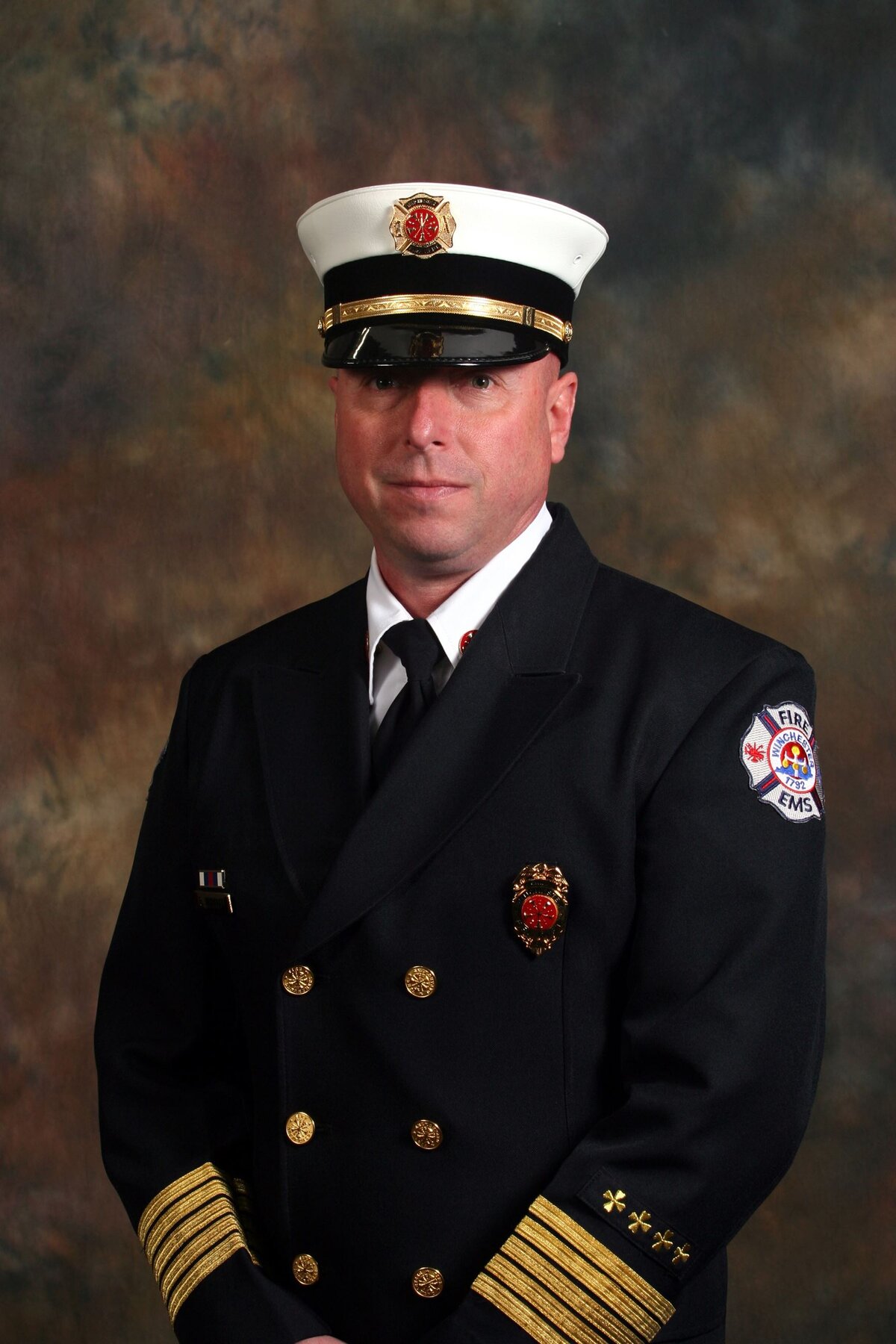 Portrait of a uniformed fire department officer wearing a formal dark dress coat with gold buttons and insignia, and a white peaked cap featuring a fire department emblem. The background is a neutral studio backdrop.