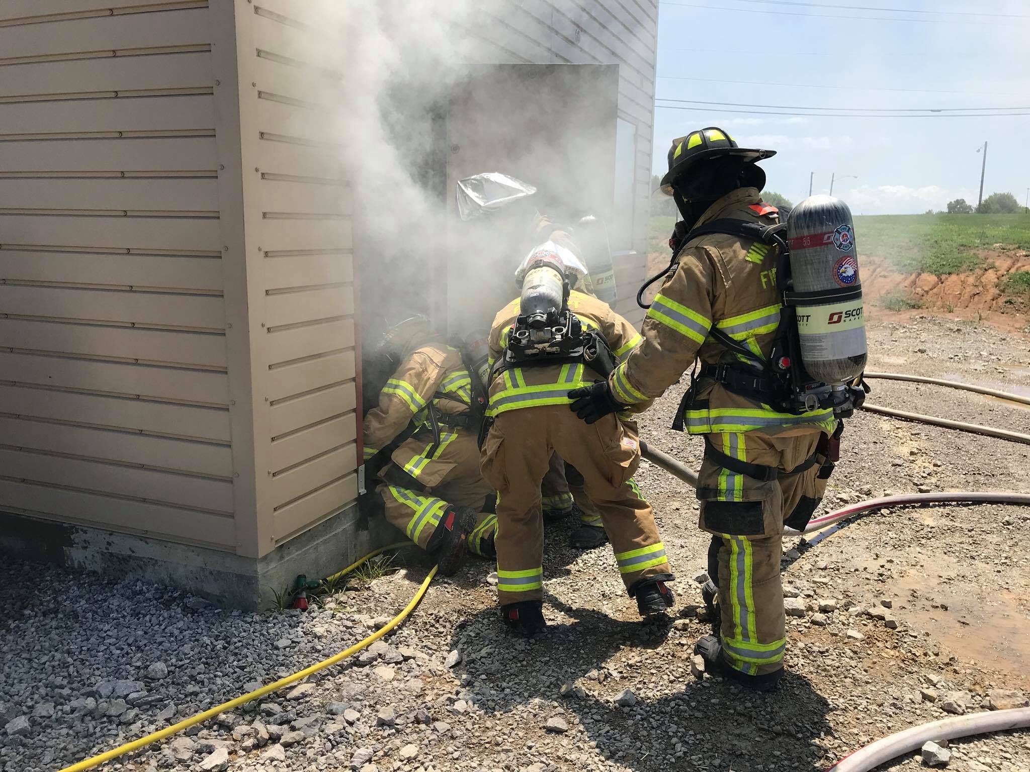 Firefighters in full protective gear and breathing apparatus are entering a smoke-filled doorway during a training exercise. Several firefighters are crouched near the entrance while another stands nearby holding a hose. The scene is outdoors next to a beige building with gravel on the ground.