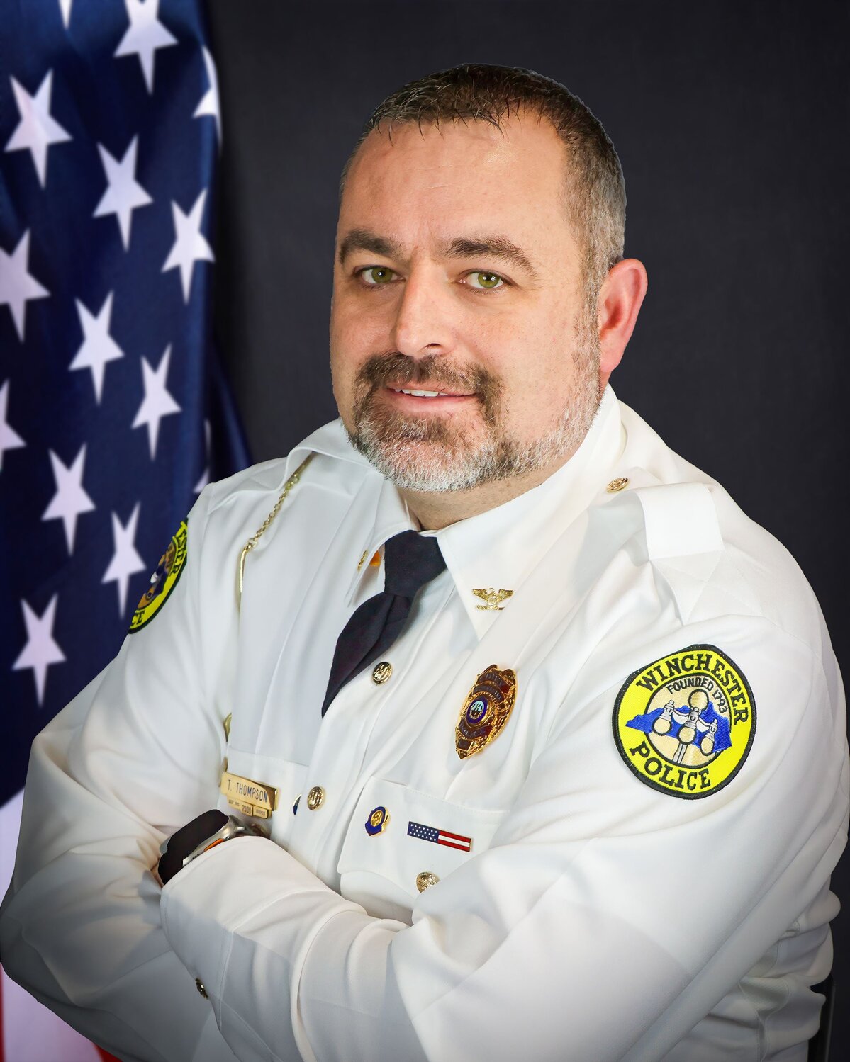 Person wearing a white police dress uniform with Winchester Police patches on the sleeves, standing in front of an American flag.