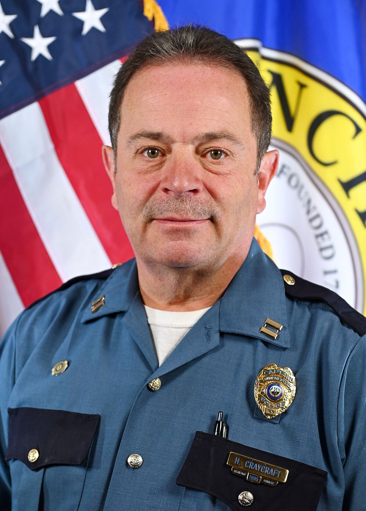 Person wearing a dark blue police uniform with visible badge and nameplate, standing in front of an American flag and a Winchester city seal banner.