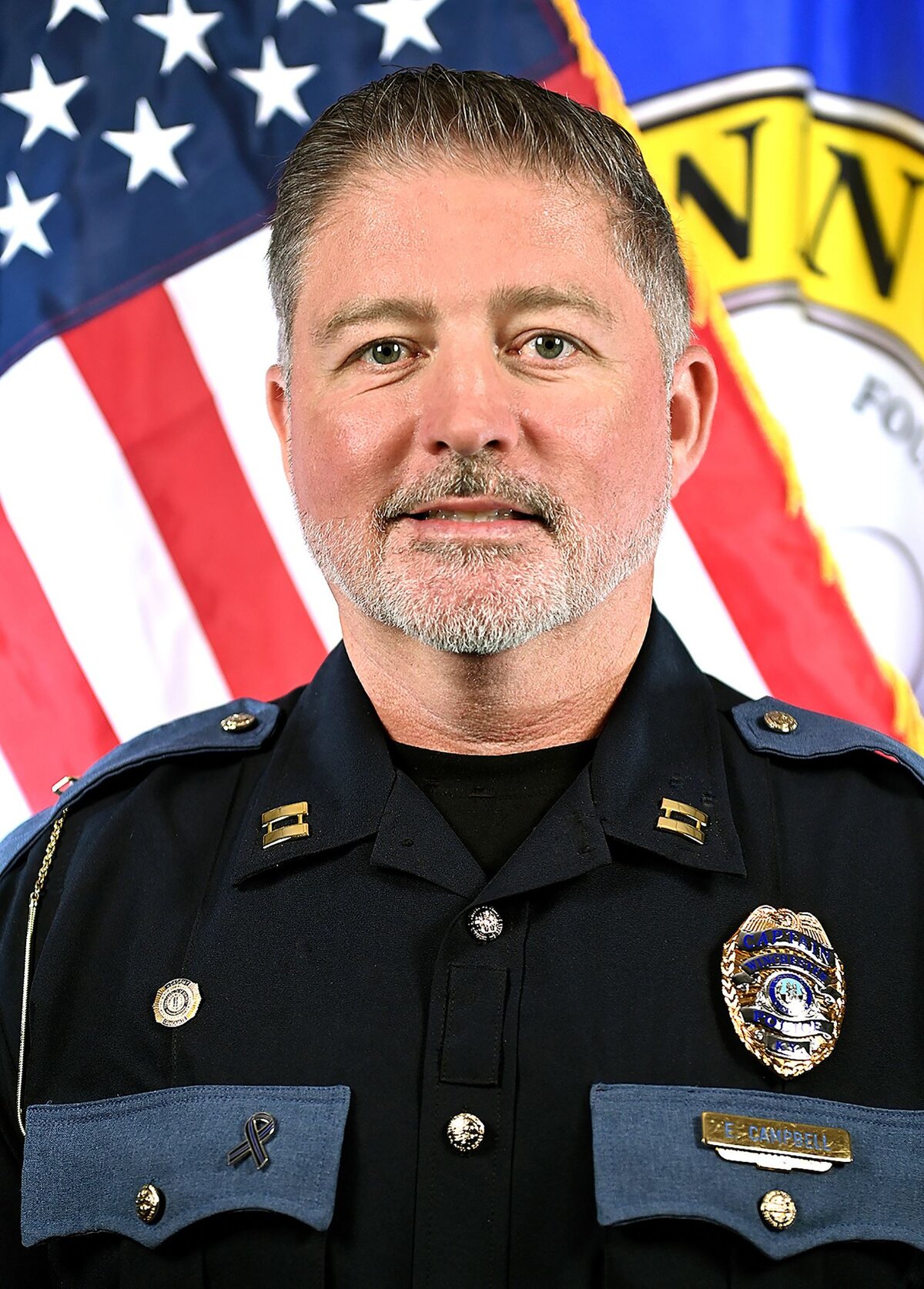 Person wearing a dark formal uniform with shoulder epaulets and gold pins. The uniform includes a badge and a nameplate that reads “E. Edwards.” A ribbon pin is visible on the left pocket flap. In the background, there is an American flag and part of a circular emblem with visible text “Winchester” and “Founded 1793.”