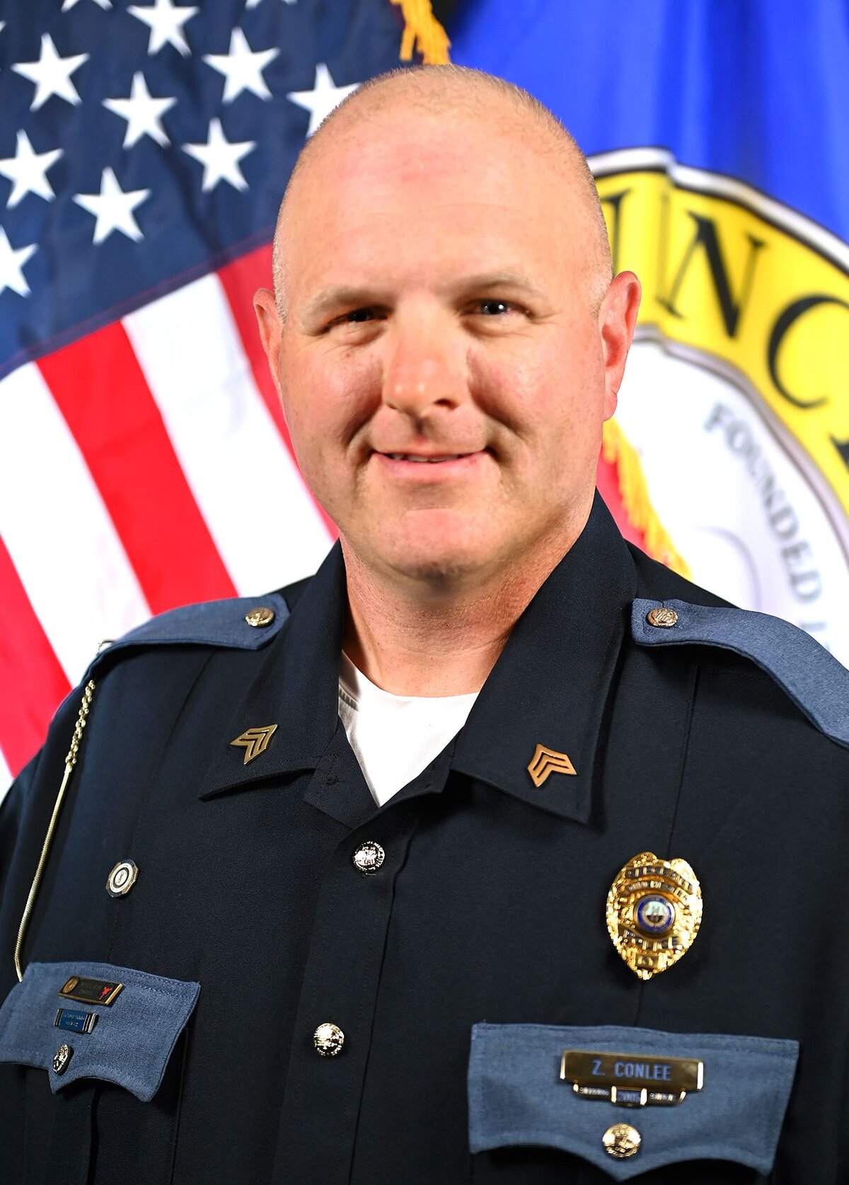 Person wearing a dark formal uniform with shoulder epaulets and a badge. The uniform includes a nameplate reading “Z. Conlee” and gold chevrons on the collar. In the background, there is an American flag and part of a circular emblem with visible text “Winchester” and “Founded 1793.”