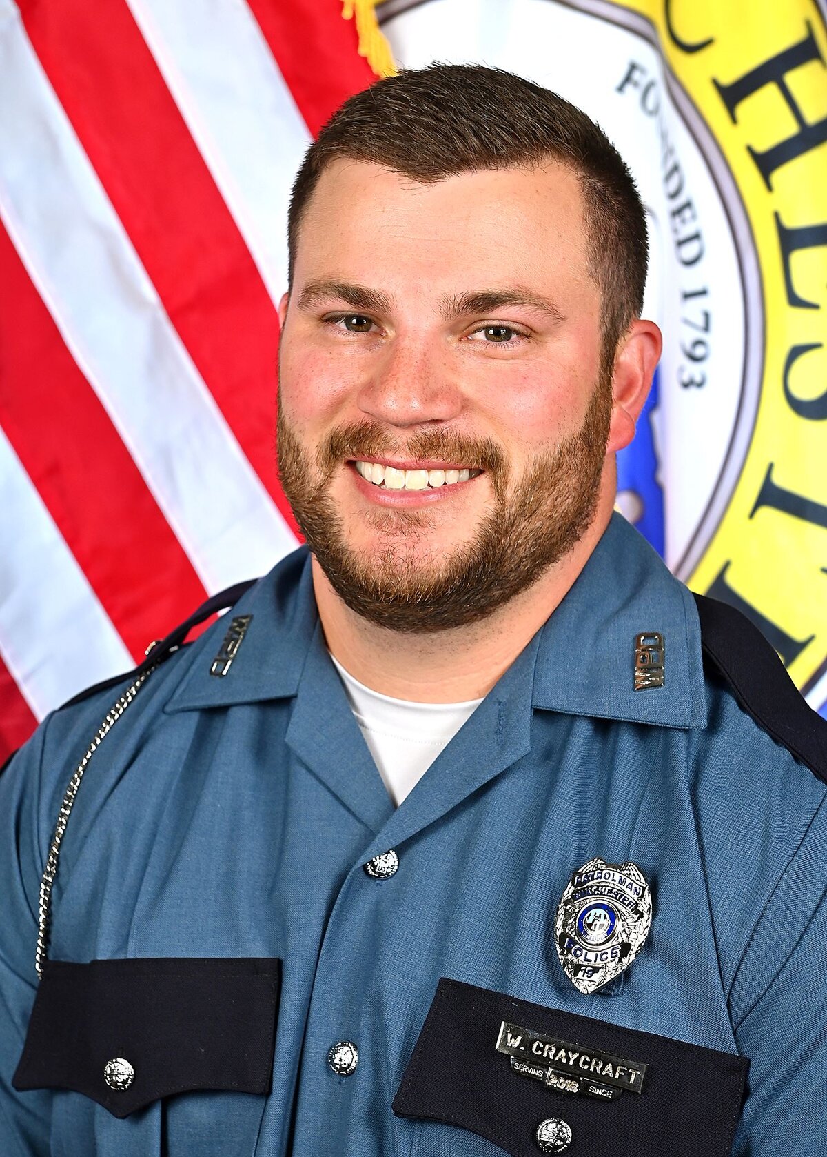 Person wearing a blue formal uniform with black accents and silver buttons. The uniform includes a badge, shoulder epaulets, and a nameplate reading “W. Craycraft.” An American flag and part of a circular emblem with visible text “Winchester” and “Founded 1793” are in the background.