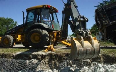 Yellow backhoe loader with a large bucket digging into rocky soil at a construction site. A dump truck is partially visible nearby.
