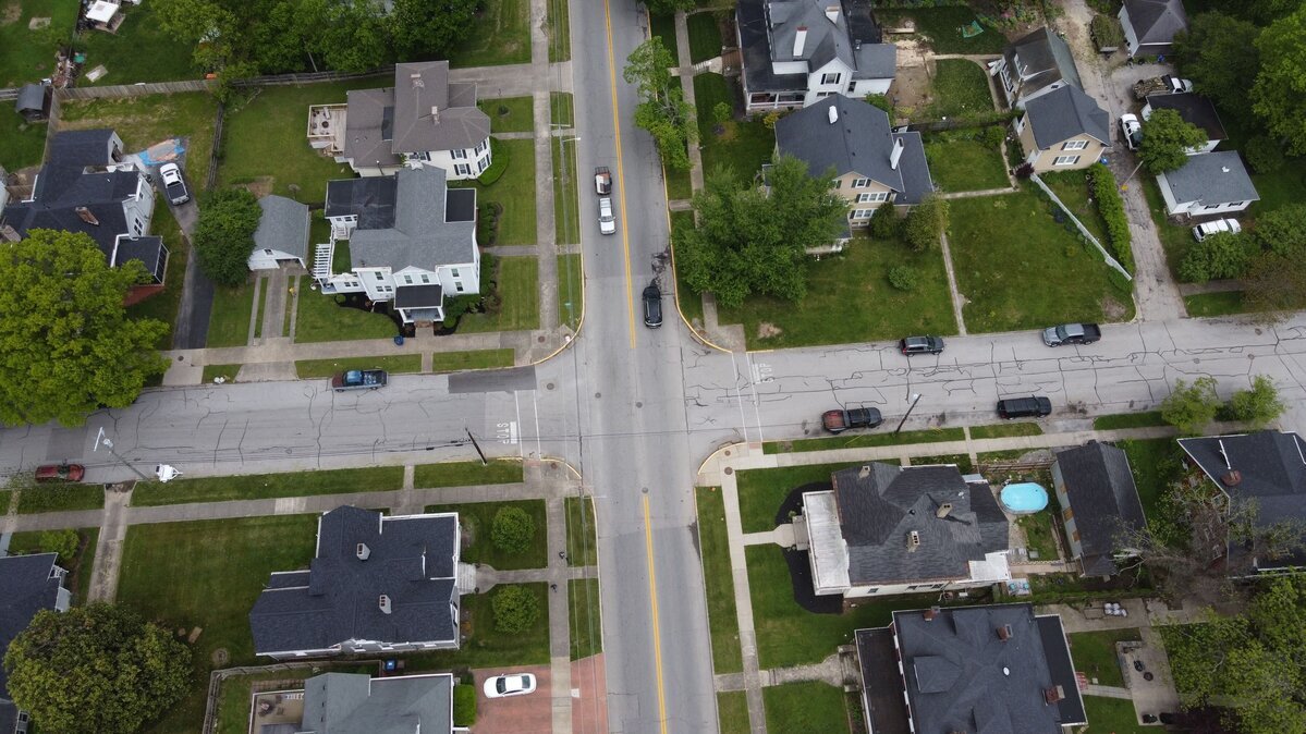 Aerial view of a residential neighborhood showing an intersection of two two-lane streets. Houses with driveways and green lawns line both sides of the streets. Several vehicles are parked along the curbs, and one vehicle is driving through the intersection.