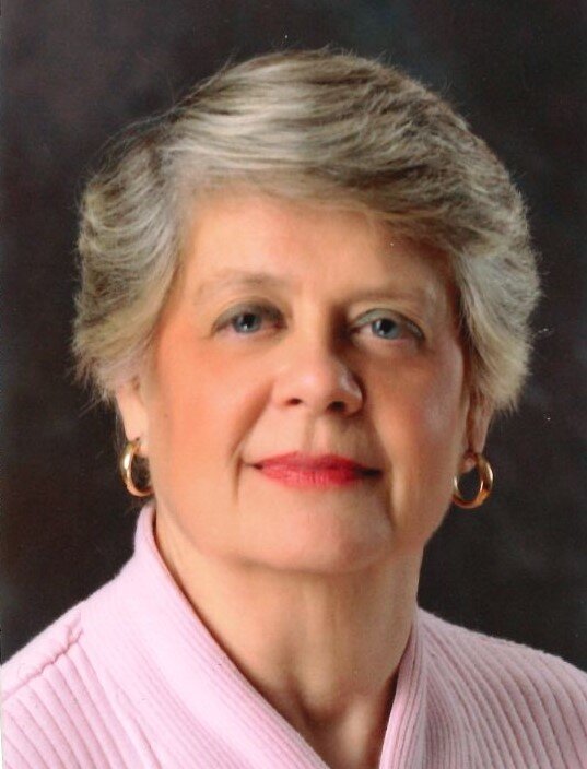 Portrait of an individual wearing a light tan jacket with a rounded collar and button detail. The person has short, neatly styled hair and is wearing hoop earrings. The background is a neutral gradient.